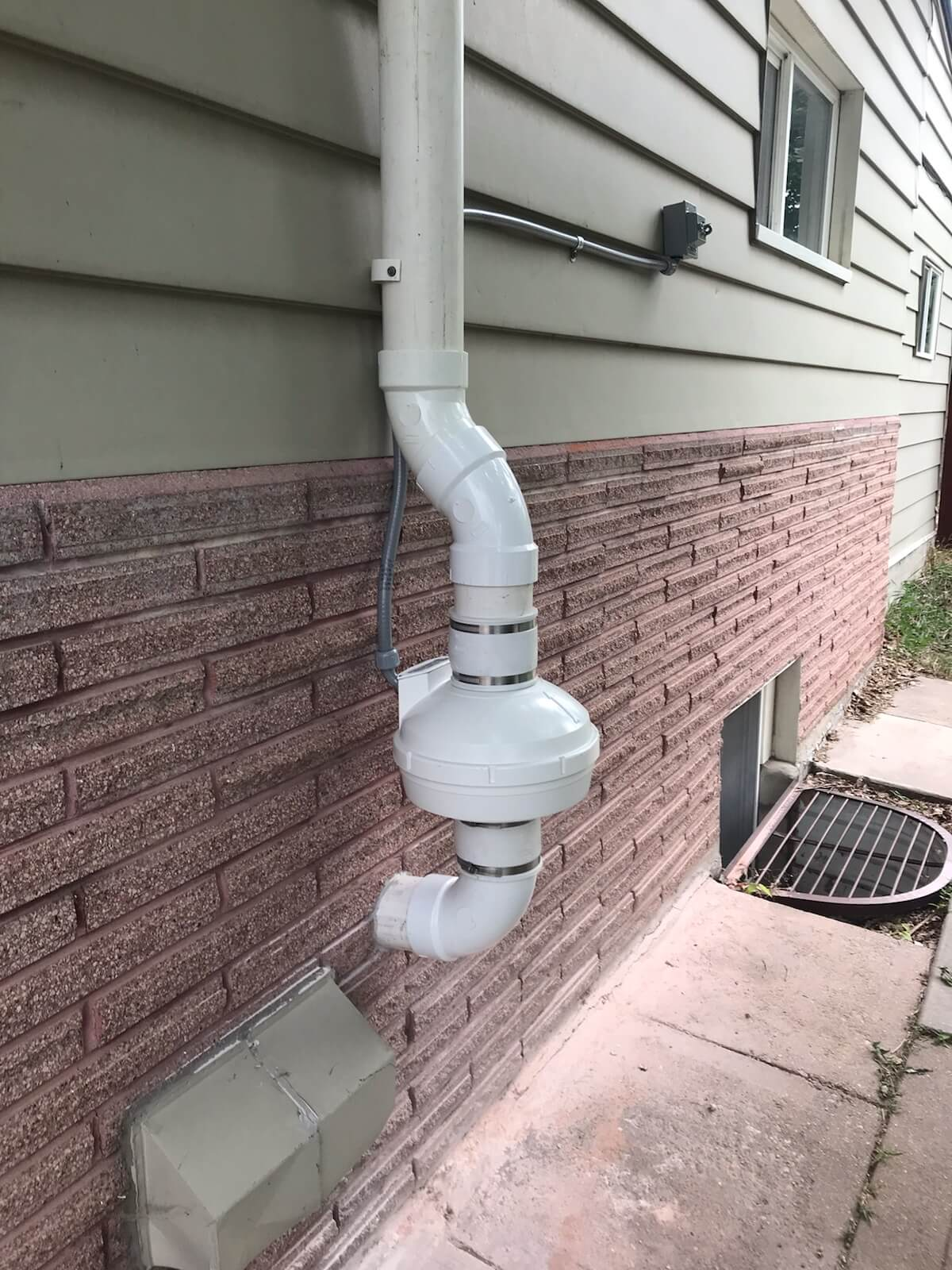 Exterior radon mitigation system fan and PVC vent stack on a residential brick foundation